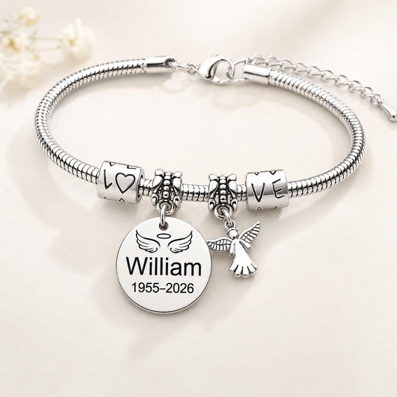 May include: A silver bracelet with several charms. The bracelet features a round charm engraved with the name "William" and the years "1955-2026". Other charms include a heart, the word "LOVE", and an angel. The bracelet is displayed on a white background.