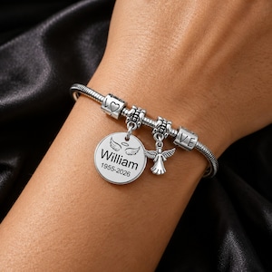 May include: Silver charm bracelet with a round pendant engraved with the name "William", angel wings, and the years 1955-2026. The bracelet also features heart and "LOVE" charms, and an angel charm.
