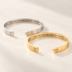 May include: Two open cuff bracelets, one in silver-tone and one in gold-tone metal. Each bracelet features the engraved text FUCK THIS, FUCK THAT, FUCK YOU, FUCK OFF along the inner band and a small black heart outline at each rounded end.