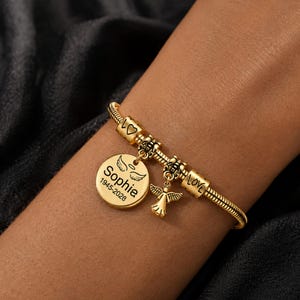 May include: Gold charm bracelet with a round pendant engraved with the name "Sophie", angel wings, and dates 1945-2028. The bracelet also features a small angel charm and charms that spell "LOVE".