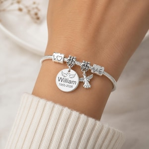 May include: A silver charm bracelet featuring several charms. The bracelet includes a round charm engraved with the name "William" and the years "1955-2026", an angel charm, and charms with hearts and the word "LOVE".