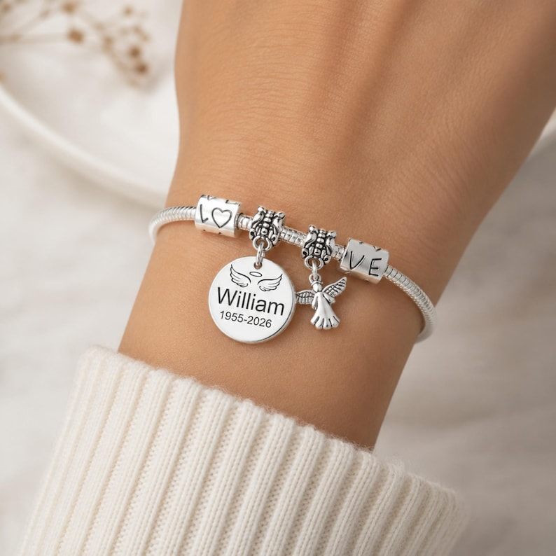 May include: A silver charm bracelet featuring several charms. The bracelet includes a round charm engraved with the name "William" and the years "1955-2026", an angel charm, and charms with hearts and the word "LOVE".
