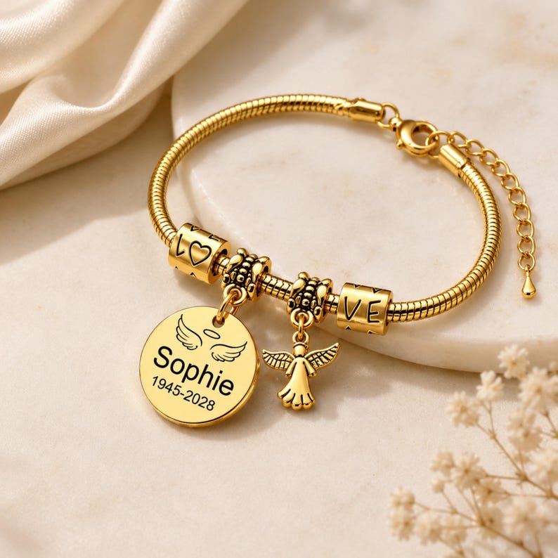 May include: Gold-coloured bracelet with a snake chain. It has charms spelling "LOVE", a heart, and a round pendant engraved with "Sophie" and dates. An angel charm hangs from the bracelet.