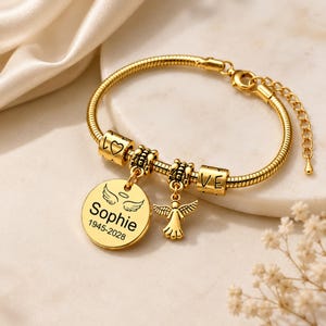May include: Gold-coloured bracelet with a snake chain. It has charms spelling "LOVE", a heart, and a round pendant engraved with "Sophie" and dates. An angel charm hangs from the bracelet.