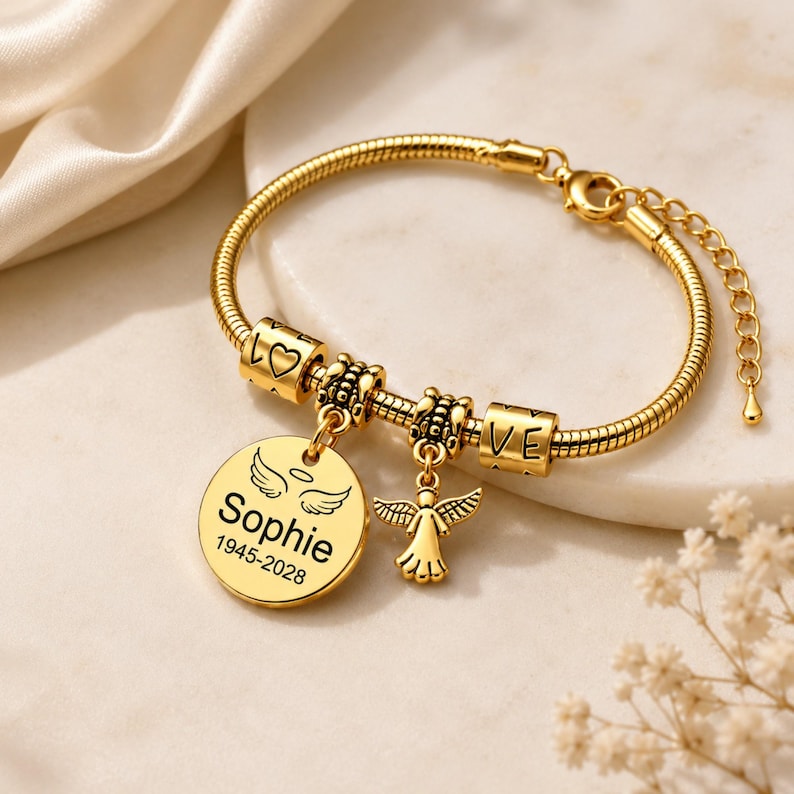 May include: Gold-coloured bracelet with a snake chain. It has charms spelling "LOVE", a heart, and a round pendant engraved with "Sophie" and dates. An angel charm hangs from the bracelet.