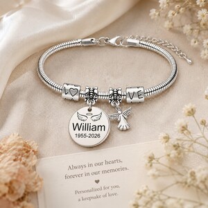 May include: Silver charm bracelet featuring a round pendant engraved with "William" and dates "1955-2026". The bracelet includes charms with the words "LOVE" and an angel charm. The bracelet is displayed on a cream-coloured surface.