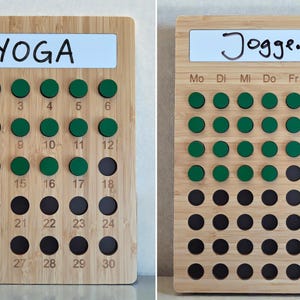 Könnte beinhalten: Zwei rechteckige Gewohnheits-Tracker aus Bambus mit weißen Whiteboard-Flächen oben. Das linke Brett zeigt den Text YOGA und die Zahlen 1 bis 30 mit grünen magnetischen Chips auf den Zahlen 1 bis 18. Das rechte Brett trägt die Aufschrift Joggen.