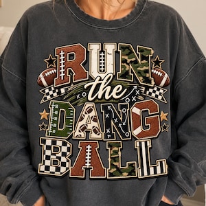 May include: A charcoal grey crewneck sweatshirt featuring a graphic print with the text RUN the DANG BALL. The design incorporates football-themed patterns, including brown football textures, green camouflage, and black and white checkerboard.