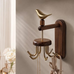 May include: A wall-mounted jewellery organiser featuring a dark wood vertical base and horizontal arm. A gold-toned metal bird sits atop the arm, connected to a rotating circular wood platform with six curved gold hooks for hanging necklaces and pouches.