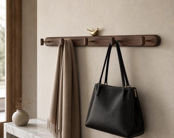 May include: A horizontal dark wood wall-mounted coat rack featuring five pegs and a small gold-toned bird figurine perched on top. A beige scarf and a black leather handbag with two shoulder straps are hanging from the wooden pegs.