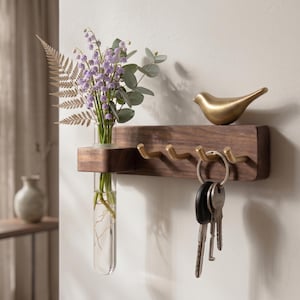 May include: A wooden key holder with brass hooks and a small shelf. A clear glass tube vase holds purple flowers and greenery. A gold-colored bird figurine sits on the shelf. Keys hang from one of the hooks.