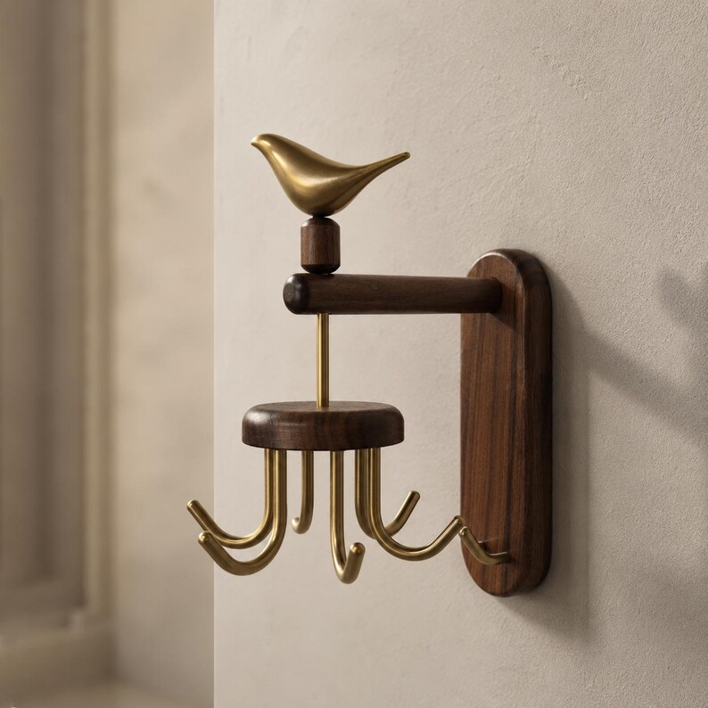 May include: A wall-mounted hook rack featuring a dark brown wooden base and horizontal arm. A gold-toned metal bird sits atop the arm. Below, a circular wooden platform supports four curved gold-toned metal hooks for hanging items securely.