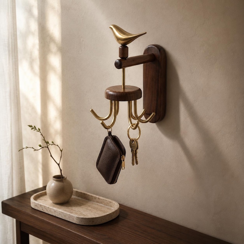 May include: A wall-mounted key holder featuring a dark wood rectangular base and a horizontal arm. A brass bird figurine sits atop the arm, which supports a rotating circular wooden platform with four brass hooks holding a leather pouch and keys.