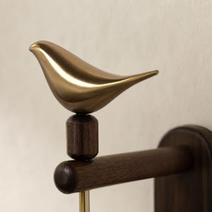 May include: A wall-mounted fixture featuring a smooth, metallic gold-toned bird sculpture perched atop a dark brown wooden cylindrical post. The post is attached to a horizontal wooden arm, which connects to a vertical wooden wall-mounting plate.