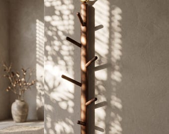 May include: A wooden wall-mounted coat rack with multiple pegs, resembling a tree trunk. It is made of dark brown wood and topped with a small, gold-colored bird. The rack is mounted on a light-colored wall, with sunlight casting shadows.