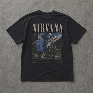 Op de afbeelding: Zwart t-shirt met de bandnaam "NIRVANA" in een verweerde lettertype boven een afbeelding van een engel met vleugels. Het ontwerp bevat blauwe en beige accenten, met extra tekst en illustraties eronder.