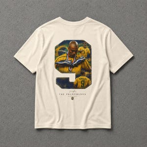 Puede incluir: Camiseta color crema con un gráfico de un jugador de fútbol con el número 9. El gráfico incluye un retrato del jugador, una bandera brasileña y el texto "THE PHENOMENON NEVER DIES."