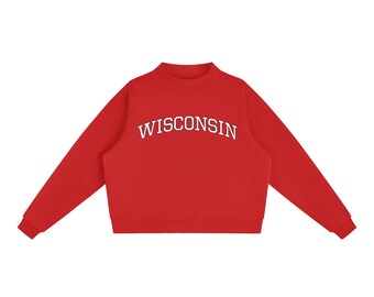 May include: A bright red long-sleeve sweatshirt featuring a mock neck collar and ribbed cuffs. The word WISCONSIN is printed in a white, arched, serif font across the center of the chest. The garment has a straight hemline.