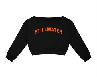 May include: A black long-sleeve cropped sweatshirt featuring the word STILLWATER printed in a bold, arched orange font across the chest. The garment has a wide neckline, cuffed sleeves, and a straight hemline at the waist.