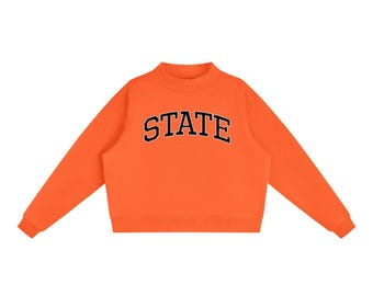May include: A bright orange long-sleeve sweatshirt featuring a mock neck collar and ribbed cuffs. The word STATE is printed across the chest in a bold, arched black font with a white outline, creating a clear contrast against the orange fabric.