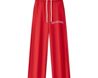 May include: Bright red sweatpants featuring a wide-leg silhouette, an elastic waistband with long white drawstring cords, and the word OKLAHOMA printed in white capital letters on the upper right leg area.