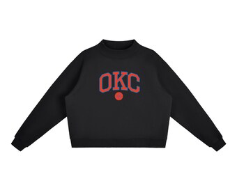 May include: A black long-sleeve sweatshirt featuring a mock neck collar and ribbed cuffs. The chest displays the text OKC in a bold, arched orange font with a blue outline, positioned above a small, solid orange circular graphic.