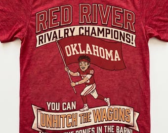May include: A red short-sleeved t-shirt featuring a graphic of a football player holding an Oklahoma flag. Text reads: RED RIVER RIVALRY CHAMPIONS! OKLAHOMA, YOU CAN UNHITCH THE WAGONS AND PUT THE PONIES IN THE BARN! OCTOBER 10, 2028.
