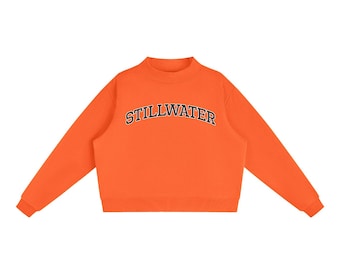 May include: A bright orange long-sleeve sweatshirt featuring a mock neck collar and ribbed cuffs. The word STILLWATER is printed in a dark navy blue collegiate-style font across the chest in a slight arch, creating a bold contrast.