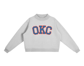 May include: A light gray long-sleeve sweatshirt with a mock neck collar and ribbed cuffs. The center of the chest features the letters OKC in a bold, collegiate-style font with blue lettering and a thick orange outline.
