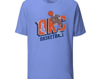 May include: A light blue short-sleeve t-shirt featuring a graphic of a brown bison mascot dunking an orange basketball. The text OKC is printed in large orange letters with a blue lightning bolt, and the word BASKETBALL is below in dark blue.
