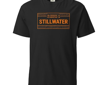 May include: A black short-sleeve t-shirt featuring an orange rectangular graphic on the chest. The graphic contains the text STILLWATER, coordinates 36.1305210 N -97.0839725 W, and a topographic line pattern within the border of the rectangle.