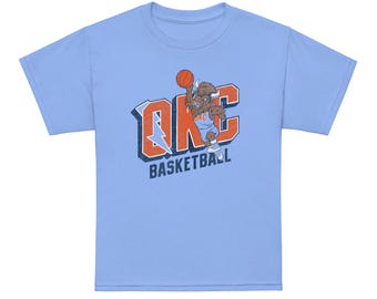 May include: A light blue short-sleeve t-shirt featuring a graphic of a brown bison wearing a basketball jersey and holding a basketball. The text OKC is displayed in large orange letters with a blue lightning bolt, and the word BASKETBALL is below.