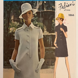 May include: A vintage Vogue Couturier Design fashion magazine cover featuring a woman in a light blue dress and hat. The cover also includes a drawing of a woman in a black dress and yellow hat. The text "Fabiani of Italy 1866" is visible.