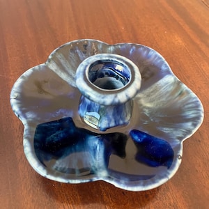 May include: A ceramic candle holder shaped like a five-petaled flower with a glossy glaze in shades of deep navy blue, royal blue, and white. The center features a raised circular socket for a taper candle, with a smooth, reflective finish.