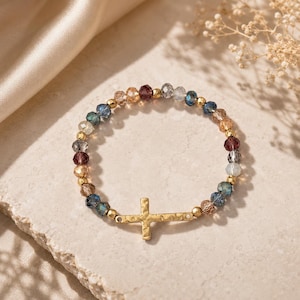 May include: A beaded bracelet featuring a gold cross charm. The bracelet is made of colorful, faceted beads in shades of blue, purple, gold, and clear. The cross is a simple, textured design.