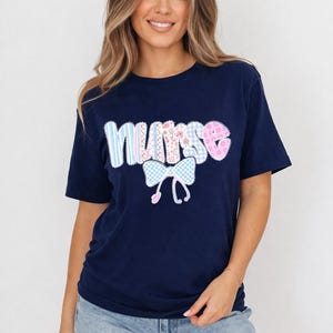 May include: Navy blue t-shirt with the word "nurse" in a patchwork design. The letters are in various patterns, including stripes, gingham, and floral prints. A light blue gingham bow is below the word.