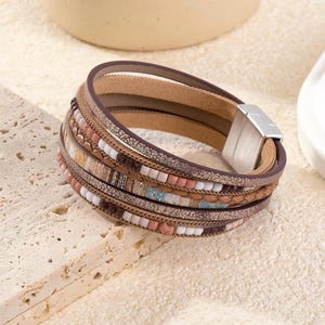 May include: A multi-layered bracelet with a magnetic clasp. The bracelet features various textures and colors, including brown, tan, and metallic gold. Accents include small beads in shades of pink, white, and blue. The bracelet is approximately 7 inches in circumference.