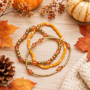 May include: A set of three beaded bracelets featuring a mix of metallic gold spheres, translucent orange faceted crystals, and small opaque yellow beads. The bracelets are arranged in a circular pattern, showcasing a variety of textures and colors.