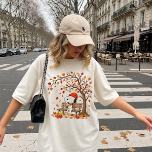 Könnte beinhalten: Ein cremefarbenes T-Shirt mit einer Grafik von Snoopy, der eine orangefarbene Mütze und einen Schal trägt und auf einer Bank unter einem Herbstbaum mit fallenden Blättern sitzt. Dazu eine beigefarbene Baseballkappe und eine schwarze Stepptasche.
