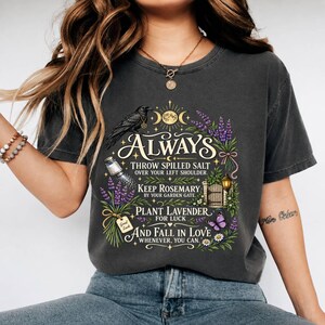 May include: A charcoal gray t-shirt featuring a graphic print with a black raven, a salt shaker, lavender sprigs, a garden gate, and a butterfly. The text reads: ALWAYS THROW SPILLED SALT OVER YOUR LEFT SHOULDER. KEEP ROSEMARY BY YOUR GARDEN GATE. PLANT LAVENDER FOR LUCK AND FALL IN LOVE WHENEVER, YOU CAN.