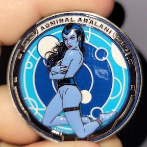 Könnte beinhalten: Eine runde, silberfarbene Münze mit einer blauhäutigen Frau mit schwarzen Haaren und roten Lippen. Die Münze trägt den Text "ADMIRAL ARALANI" am Rand und ein blau-weißes Design in der Mitte. Die Frau kniet mit verschränkten Armen.