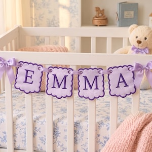 May include: A decorative banner featuring four scalloped, light purple striped fabric squares, each embroidered with a dark purple letter to spell EMMA. The banner is strung on a light purple ribbon and tied to a white crib with matching bows.