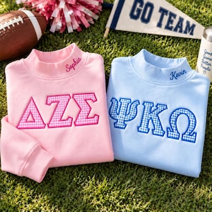 May include: A light pink sweatshirt with the Greek letters ΔΖΣ and the name Sophia embroidered in pink, and a light blue sweatshirt with the Greek letters ΨΚΩ and the name Kevin embroidered in blue, both featuring a gingham pattern.