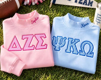 May include: A light pink sweatshirt with the Greek letters ΔΖΣ and the name Sophia embroidered in pink, and a light blue sweatshirt with the Greek letters ΨΚΩ and the name Kevin embroidered in blue, both featuring a gingham pattern.