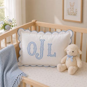 May include: A white rectangular pillow with a scalloped blue edge and a blue and white gingham monogram featuring the letters O, J, and L. The pillow is positioned in a wooden crib next to a cream-colored plush lamb toy wearing a blue gingham bow.