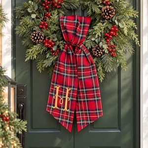 May include: A holiday wreath made of green pine needles and cedar branches, decorated with red berries and brown pinecones. A long red, green, and blue plaid fabric ribbon hangs from the center, featuring a gold embroidered letter H.