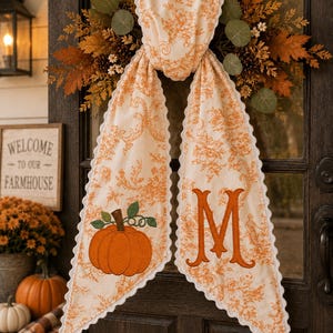 May include: A long, cream-colored fabric sash with an orange floral toile pattern and scalloped edges. One end features an embroidered orange pumpkin with green leaves, while the other end displays a large, embroidered orange letter M.