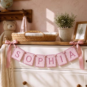 May include: A decorative banner featuring six scalloped, light pink and white striped fabric pennants, each embroidered with a dark pink letter to spell the name SOPHIA. The banner is finished with light pink satin ribbons on both ends.