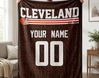 May include: A dark brown fleece blanket featuring the text CLEVELAND in large white letters, a football helmet icon, the text EST. 1946, the placeholder text YOUR NAME, and the number 00 in white, with a repeating CLEVELAND pattern in red outline.