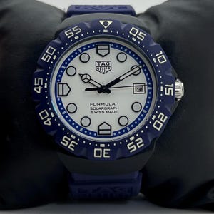 May include: A TAG Heuer Formula 1 Solargraph watch with a white dial, black hands, and circular hour markers. The watch features a blue rotating bezel with white numerals, a date window at the three o'clock position, and a blue rubber strap.